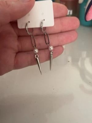Silvriga örhängen med pärldetalj och spike - Säljer ett par trendiga örhängen i silverfärg med kedjelänk, vit pärldetalj och spetsig spike längst ner. Perfekta för dig som gillar en edgy stil med en touch av elegans. Materialet ser ut att vara metall och pärlimitation.