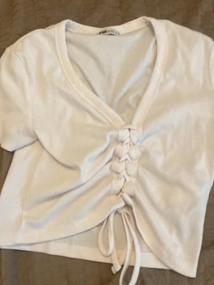 Vit croppad topp med snörning - Trendig vit croppad topp med korta ärmar och snygg snörning framtill. Toppen har v-ringning och är tillverkad i ett mjukt, ribbat material som ger en bekväm känsla och snygg passform. Perfekt för att lyfta din outfit med en cool detalj.