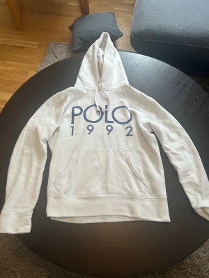Ralph lauren hoodie 1992 vit - Snygg vit hoodie med stor huva och dragsko. Framtill finns ett stort blått POLO 1992-tryck och en klassisk magficka. Hoodien har ribbade muddar vid ärmslut och nederkant. Perfekt för en avslappnad och trendig stil.