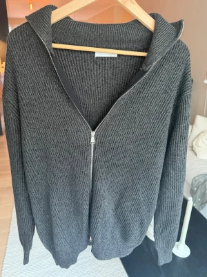 Mörkgrå ribbad kofta från Zara - Snygg mörkgrå ribbad kofta från Zara med dragkedja framtill och hög krage. Koftan är långärmad och har en relaxed passform som passar perfekt till lager på lager. Materialet är stickat och känns mjukt mot huden.
