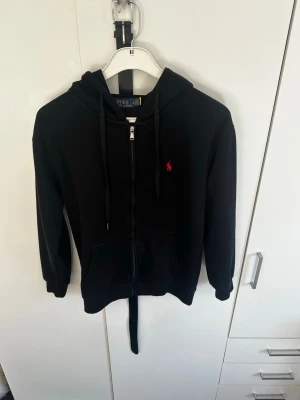 Svart hoodie med dragkedja från Polo Ralph Lauren - Säljer en svart hoodie från Polo Ralph Lauren med dragkedja framtill och röd broderad logga på bröstet. Hoodien har huva med dragsko, två fickor fram och ribbade muddar vid ärmar och nederkant. Perfekt för en chill och stilren look.