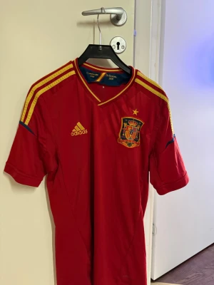 Spaniens landslagströja Adidas röd - Röd fotbollströja från Adidas med Spaniens landslagsemblem på bröstet och gula detaljer på axlarna. Tröjan har korta ärmar, V-ringning och är tillverkad i ett lätt och ventilerande material. Perfekt för fotbollsträning eller att bära när du hejar på Spanien.