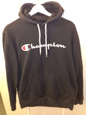 Svart Champion hoodie med logga - Snygg svart hoodie från Champion med stor vit logga och röd detalj på bröstet. Klassisk känguruficka framtill och vita dragsnören i huvan. Perfekt för en avslappnad och sportig stil. Tillverkad i mjukt material för extra komfort.