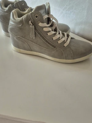 Gråa wedge sneakers med dragkedja - Snygga gråa sneakers med kilklack och vita snören. Skorna har dragkedja på sidan och perforerade detaljer för extra stil. Tillverkade i ett matt syntetmaterial med vit sula. Perfekta för dig som vill ha lite höjd men ändå sneakers-look.Oanvända, endast testade.Stl:39
