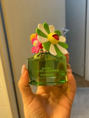 Daisy Marc Jacobs Murakami parfym - Unik parfym från Marc Jacobs i samarbete med Murakami. Flaskan är grön och har en cool, färgglad blomma som lock med vita, gröna och rosa kronblad. Designen är lekfull och sticker ut på hyllan. Perfekt för dig som gillar snygga och kreativa parfymflaskor.