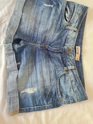 Blå denimshorts med slitningar - Snygga blå denimshorts med slitna detaljer och uppvikta benslut. Klassisk femficksmodell med knapp och dragkedja framtill. Perfekta för varma dagar och ger en avslappnad look.