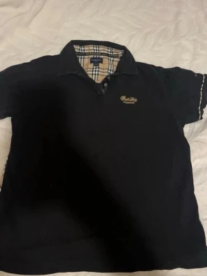 Vintage burberry tshirt - Säljer en svart äkta burberry tshirt med klassisk krage och rutiga detaljer på insidan av kragen och längs ärmarna. Det står att Tshirten är storlek L men den skulle passa utmärkt som en S/M. Tshirten har knappar framtill och en liten broderad logga på bröstet. Perfekt för en stilren och avslappnad look.