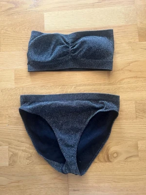 Glittrig bandeau bikini i svart - Säljer en snygg svart bandeau bikini med glittrigt tyg. Överdelen är axelbandslös med lätt rynkning framtill och underdelen har klassisk passform. Perfekt för dig som vill sticka ut på stranden i sommar! Aldrig Andvänd, Liten i bysten 