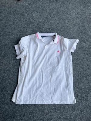 Vit pikétröja med rosa detaljer Adidas - Snygg vit pikétröja från Adidas med korta ärmar och rosa detaljer på kragen samt rosa logga på bröstet. Tillverkad i ett mjukt och sportigt material som passar perfekt för tennis eller träning. Klassisk pikékrage och knappslå framtill.