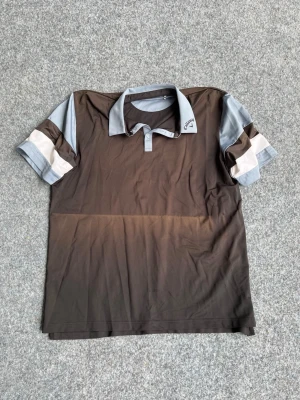 Svart och grå piké från Callaway - Snygg pikétröja från Callaway i svart och grått med ljusgrå krage och knappar. Ärmarna har vita och mörkare grå detaljer som ger en sportig look. Perfekt för golf eller andra sportaktiviteter. Tillverkad i ett lätt och svalt syntetmaterial.