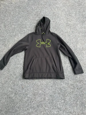 Svart Under Armour hoodie - Svart hoodie från Under Armour med stor neongul logga på bröstet och matchande text bak. Klassisk känguruficka framtill och huva med dragsko. Tillverkad i mjukt och bekvämt material, perfekt för träning eller chill.