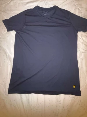 Lyle&Scott t-shirt med gul brodyr - Snygg&Lyle Scott mörkblå t-shirt i bomull med rund hals och korta ärmar. På nedre delen finns en liten gul broderad logga som ger en clean och stilren look. Perfekt till jeans eller shorts för en enkel och fräsch outfit.