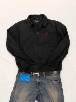 Svart bomberjacka från Polo Ralph Lauren - Svart bomberjacka från Polo Ralph Lauren med klassisk krage, dragkedja framtill och röd broderad logga på bröstet. Jackan har elastiska muddar och är tillverkad i bomull. Perfekt för en stilren och avslappnad look.