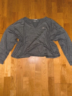 Grå croppad tröja från Hollister - Mysig grå långärmad tröja från Hollister med croppad passform och v-ringning. Tröjan har en liten logga på bröstet och är tillverkad i mjukt material som känns skönt mot huden. Perfekt för en avslappnad och trendig stil.