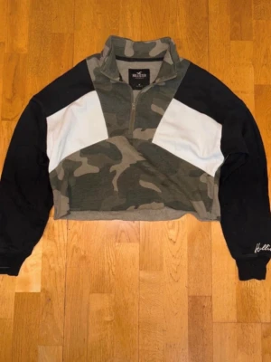 Camo  från Hollister - Snygg croppad  från Hollister med camomönster, svarta ärmar och vita detaljer framtill. Tröjan har halv dragkedja och hög krage. Perfekt för en chill och trendig look.