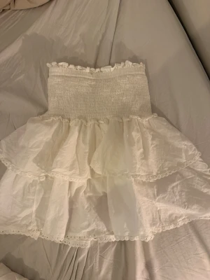 Vit volangkjol från Drôle de Copine - Supersöt vit kjol från Drôle de Copine med bred smock i midjan och dubbla volanger. Kjolen har romantiska spetsdetaljer längs kanterna och är perfekt för sommarens alla tillfällen. Lätt och luftig känsla.