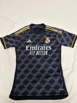 Real Madrid bortatröja Adidas - Snygg Real Madrid bortatröja i mörkgrått med grafiskt mönster och gula detaljer på axlar och ärmslut. Tröjan har klubbmärke och Adidas-logga på bröstet samt 'Emirates Fly Better' tryckt framtill. Tillverkad i ett lätt och ventilerande funktionsmaterial.