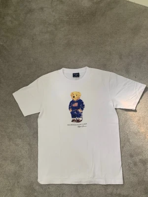 Vit t-shirt med Polo Bear tryck  - Vit t-shirt från Polo Ralph Lauren med det klassiska Polo Bear-trycket framtill. T-shirten har rund halsringning och korta ärmar.  Andvänd väldigt få gånger!!!!