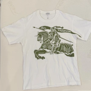 Vit Burberry t-shirt med tryck - Vit t-shirt från Burberry med stort grönt tryck av riddare och flagga med texten 'Prorsum' på framsidan. Klassisk rund halsringning och korta ärmar. Tillverkad i mjuk bomull för en bekväm känsla.