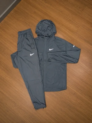 Nike grå träningsset med huva - Snyggt grått träningsset från Nike med jacka och byxor. Jackan har huva, dragkedja och reflexdetaljer på ärmarna samt Nike-logga på bröstet. Byxorna har resår i midjan och Nike-logga på låret. Perfekt för träning eller chill.
