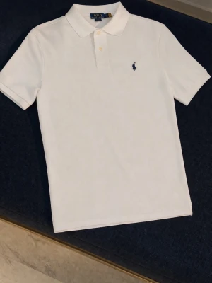 Vit pikétröja från Polo Ralph Lauren - Klassisk vit pikétröja från Polo Ralph Lauren med korta ärmar, krage och knappar framtill. Tröjan har den ikoniska broderade loggan på bröstet och är tillverkad i mjuk bomull. Perfekt för en stilren och avslappnad look.