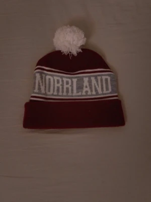 Norrlands mössa - Säljer en vinröd mössa med vit tofs och grått parti där det står NORRLAND i stora vita bokstäver. Mössan har vita och röda ränder och är stickad i mjukt material. Perfekt för kalla dagar och snygg till streetwear.