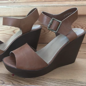 Bruna kilklackade sandaler - Oanvända. Snygga bruna sandaler med hög kilklack och breda remmar i läderimitation. Justerbart spänne vid ankeln och öppen tå. Sulan är i mörkbrunt material med mönstrad undersida för bättre grepp. Perfekta för dig som vill ha både höjd och komfort.