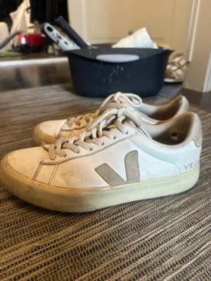 Vita Veja sneakers med beige detaljer - Snygga vita Veja sneakers med beige V-logga på sidan och matchande beige sula. Skorna har klassisk låg profil, rund tå och vita skosnören. Tillverkade i läder med textildetaljer på insidan. Perfekta för en clean och stilren look.