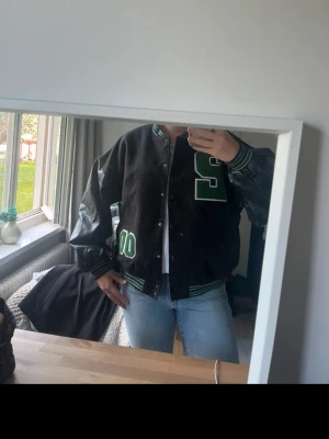 Svart bomberjacka med grön S-detalj - Säljer en svart bomberjacka i college-stil med stora gröna bokstäver och vita detaljer. Jackan har tryckknappar, ribbade muddar och krage med gröna och vita ränder. Materialet ser ut att vara en mix av tyg och skinnimitation på ärmarna. Perfekt för en sportig och trendig look.