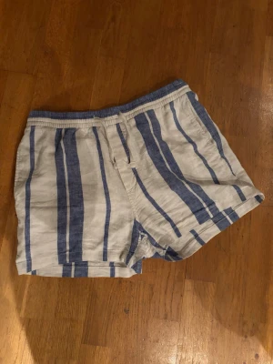 Randiga blåvita linneshorts - Snygga shorts med blå och vita ränder, elastisk midja och snörning. Tillverkade i svalt linne, perfekta för varma dagar. Shortsen har en avslappnad passform och en bakficka för extra stilpoäng.