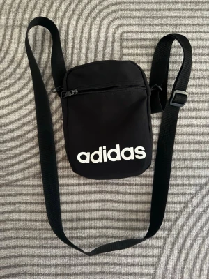 Svart axelväska från Adidas - Snygg svart axelväska från Adidas med justerbar rem och vit logga framtill. Väskan har en dragkedja upptill och är tillverkad i slitstarkt tygmaterial. Perfekt för att bära det viktigaste när du är på språng.