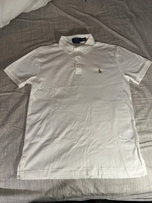 Vit pikétröja från Polo Ralph Lauren - Klassisk vit pikétröja från Polo Ralph Lauren med krage, tre knappar och den ikoniska broderade loggan på bröstet. Tillverkad i mjuk bomull och har korta ärmar. Perfekt till en clean och stilren look.