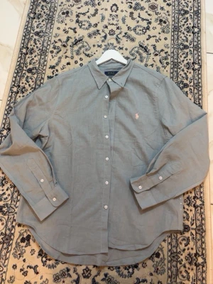 Grå skjorta från Polo Ralph Lauren - Klassisk grå skjorta från Polo Ralph Lauren med vit knappstängning och ikonisk rosa logga broderad på bröstet. Skjortan har lång ärm och en avslappnad passform, perfekt till jeans eller chinos. Tillverkad i mjuk bomull för en bekväm känsla.