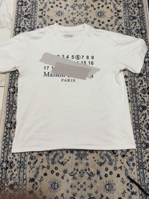 Vit Maison Margiela t-shirt - Vit t-shirt från Maison Margiela med tryck i svart på bröstet som visar siffror och texten 'Maison Margiela PARIS'. Klassisk rak passform och rund halsringning. Tillverkad i mjuk bomull för en bekväm känsla.
