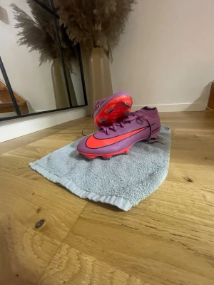 Lila Nike fotbollsskor med orange sula - Säljer ett par snygga lila fotbollsskor från Nike med en stor orange swoosh på sidan och orange sula. Skorna har klassisk snörning och är designade för spel på gräs. Perfekta för dig som vill sticka ut på planen med både stil och funktion. Finns bara i Trelleborg! Dem är använda typ i 2-3 veckor
