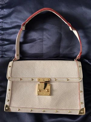 Louis Vuitton väska! Vit handväska med gulddetaljer LV  - Stilren vit handväska från Louis Vuitton i strukturerat läder med guldfärgade metalldetaljer och nitar. Väskan har ett kort handtag med röd kant och ett elegant lås framtill. Perfekt accessoar för dig som gillar lyxig och klassisk design. Sparsamt använt. Dustbag medföljer 