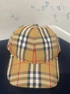 Burberry keps - Snygg keps i beige med klassiskt rutigt mönster i svart, vitt och rött. Justerbar baktill för perfekt passform. Tillverkad i bomullstyg med svart insida. Perfekt accessoar för en trendig och avslappnad stil.