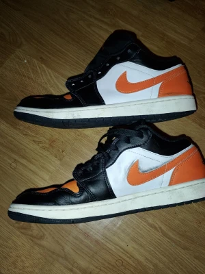 Svarta och orangea Nike Air Jordan 1 Low - Säljer ett par Nike Air Jordan 1 Low sneakers i svart, vitt och orange. Skorna har klassisk låg siluett, svart läder på ovandelen, vita paneler och orange detaljer på swoosh, häl och tå. Tjock vit sula och svart yttersula. Perfekta för dig som gillar streetwear och snygga färgkombos.