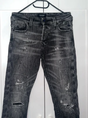 Grå slim fit slitna jeans från Jack & Jones - Säljer ett par grå jeans från Jack & Jones med slitningar, hål och distressed detaljer. Jeansen har fem fickor, bälteshällor och en klassisk knappgylf. Materialet är denim med en tvättad look. Mått 98cm 37cm skriver vid funderingar 🤩🙌