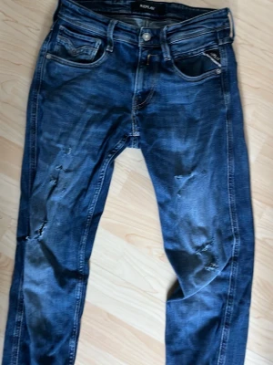 Blå slitna jeans från Replay - Säljer ett par blå jeans från Replay med slitningar och trasiga detaljer på benen. Klassisk femfickor och låg midja. Jeansen har en snygg tvättad look och kontrastsömmar. Perfekta för en avslappnad streetstil.