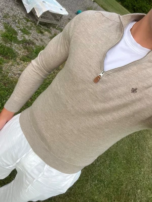 Merino Morris quarter zip - Snygg beige stickad tröja med halvzip och diskret broderad detalj på bröstet. Tröjan har en klassisk passform och ribbade muddar vid ärmslut och nederkant. Perfekt att bära över en t-shirt för en clean look. 