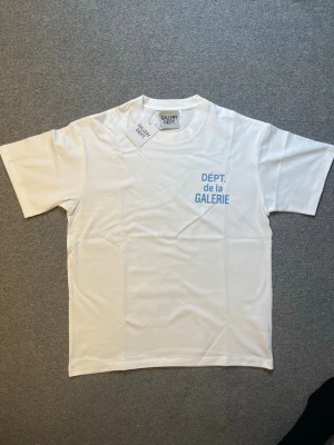 Vit t-shirt från Gallery Dept. - Vit t-shirt från Gallery Dept. med trycket 'DÉPT. de la GALERIE' i blått på bröstet. Klassisk passform med rund hals och korta ärmar. Tillverkad i mjuk bomull, perfekt för en clean och enkel stil.
