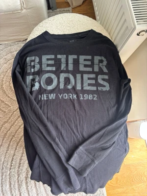 Svart långärmad topp Better Bodies - Svart långärmad topp från Better Bodies med stort tryck på bröstet där det står 'BETTER BODIES NEW YORK 1982'. Toppen har rund halsringning och är tillverkad i ett ribbat material som ger en sportig känsla.