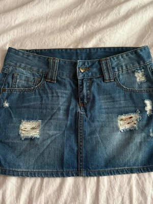 Blå jeanskjol med slitningar - Säljer en blå jeanskjol i kort modell med slitna detaljer framtill och bakfickor. Kjolen har bälteshällor, knappstängning och en liten hjärtformad detalj bak. Perfekt för dig som gillar en avslappnad och trendig stil.
