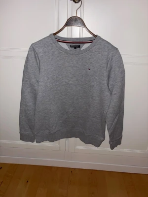 Grå sweatshirt Tommy Hilfiger - Klassisk grå sweatshirt från Tommy Hilfiger med rund halsringning och diskret logga på bröstet. Tröjan har ribbade muddar vid ärmslut och nederkant samt en enkel, stilren design som passar till det mesta.