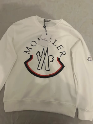 Vit Moncler hoodie med logga - Snygg vit hoodie från Moncler med stor logga framtill i svart och rött. Klassisk rund halsringning och ribbade muddar. Moncler-märke på ärmen. Perfekt för dig som gillar stilrena och exklusiva plagg.