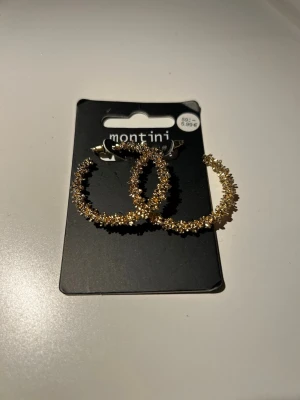 Glittriga guldiga hoops från Montini - Säljer ett par glittriga guldiga hoop-örhängen från Montini. Örhängena har en rund form och är täckta av små gnistrande detaljer som ger en extra festlig look. Perfekta för att lyfta vilken outfit som helst. Oanvända.
