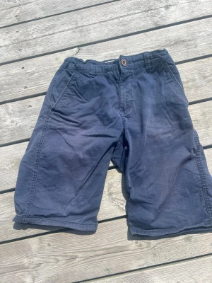 Mörkblå shorts från Radon Republic, stl 164 - Säljer ett par mörkblå shorts från Radon Republic i storlek 164. De har klassisk passform med fickor på sidorna och knappstängning i midjan. Perfekta för varma dagar och har en enkel, stilren look.
