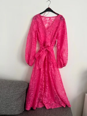 Klänning  - Säljer en lång, transparent rosa klänning med v-ringning, volangdetaljer och knytband i midjan. Klänningen har långa, puffiga ärmar och ett blommigt mönster i tyget. Perfekt för dig som vill sticka ut med en färgstark och feminin look.