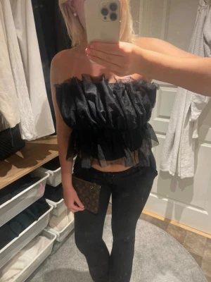 Svart volangig tubtopp - Säljer en svart tubtopp med flera lager volanger i ett transparent, lätt material. Toppen är axelbandslös och har en croppad passform som ger en edgy och trendig look. Perfekt för dig som vill sticka ut med en unik topp.